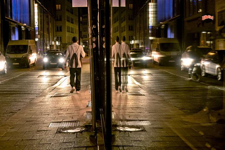 A person walking away down the street at night, cars coming in this direction, with the reflection of the same scene in a shop window splitting the image down the middle