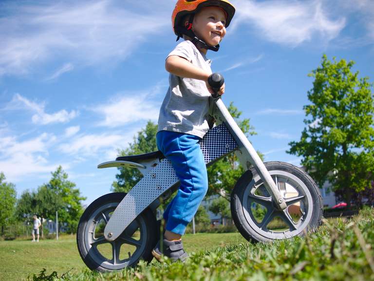 A kid on a wooden balance bike, on a green field with blue skies in the background