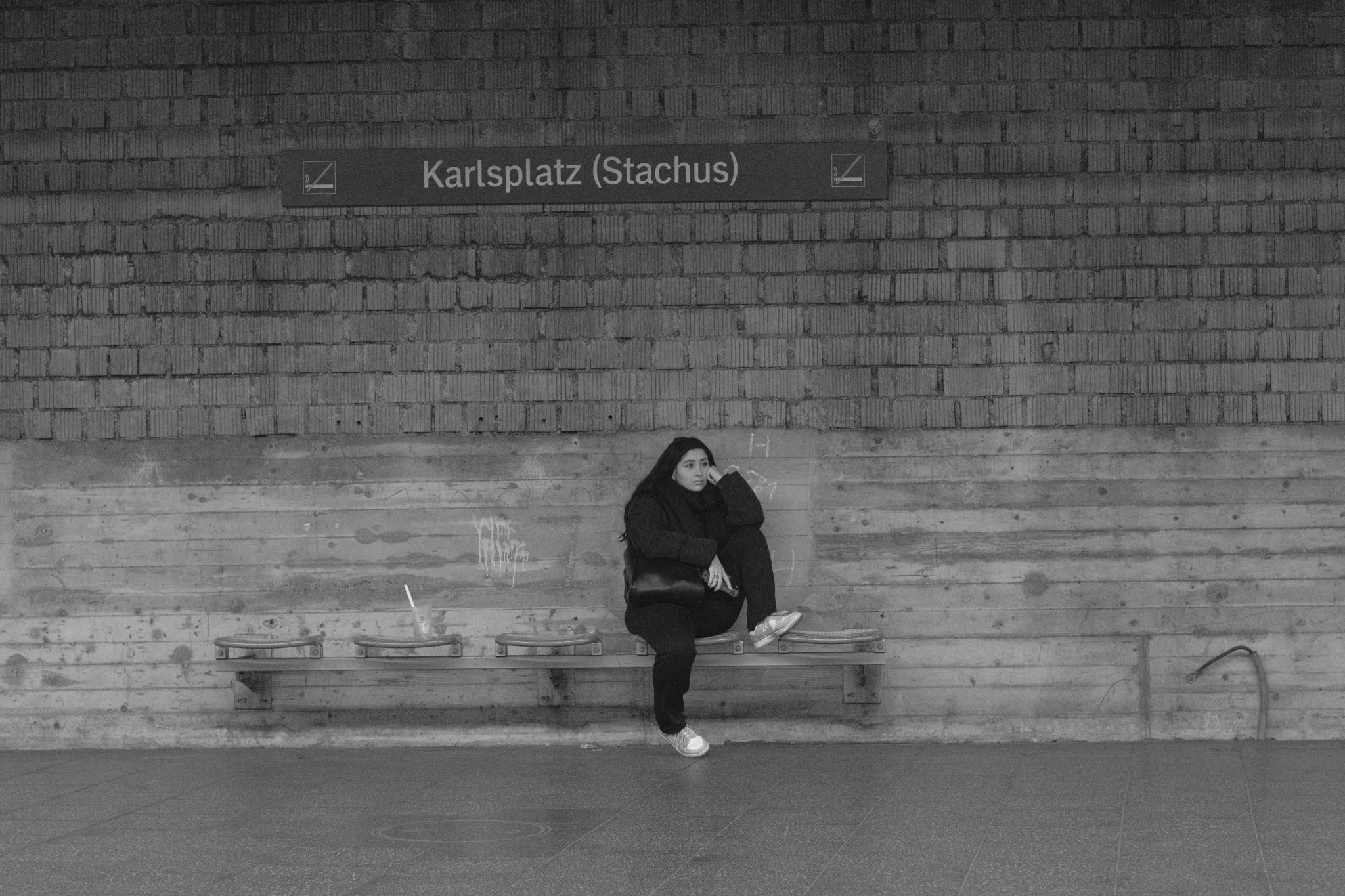 A woman sitting on a bench in front of a rough wall on a subway station. A sign above her says "Karlsplatz (Stachus)".