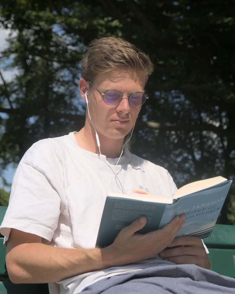 A man in a white shirt, with glasses, sitting on a bench reading a book.