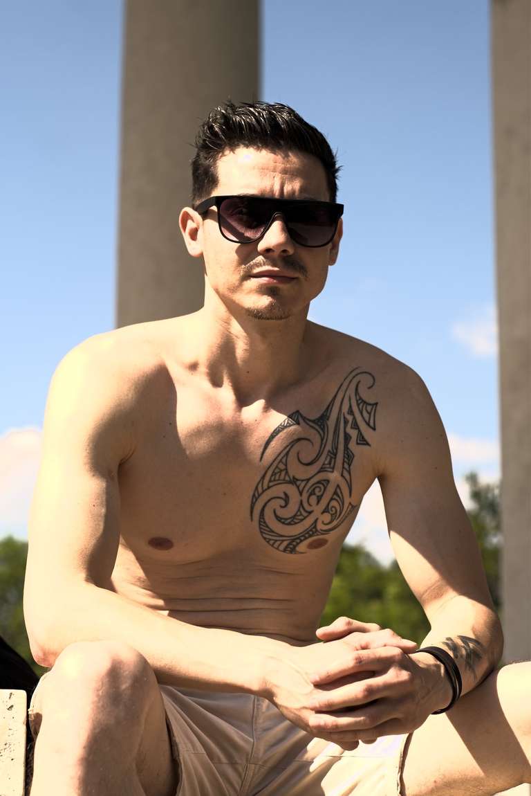 A shirtless man sitting outdoors, hands in front of him, with a tattoo going in from his shoulder into the torso